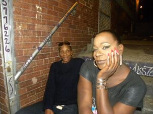 Two women posing casually in an indoor setting with a brick wall background.