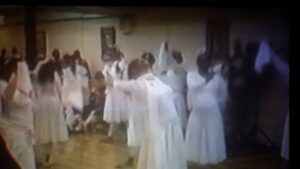 People in white robes dancing in a room with wooden floor.