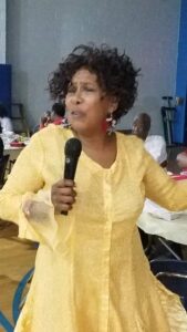 Woman speaking into a microphone, wearing a yellow blouse.