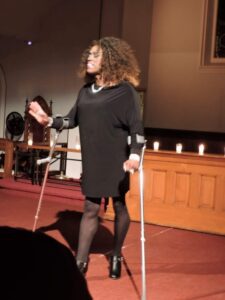 Woman with crutches speaking on stage in a dimly lit room.