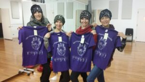 Four women holding purple scarves with a design and text, smiling indoors.