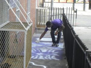 Person painting on a sidewalk near a fence.