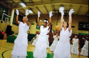 Three women in white dresses dancing indoors, each holding a white fan.