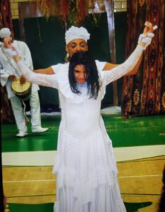 A woman in white dances joyfully with a man behind her in a lively setting.