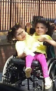 Two children smiling, one in a wheelchair, sharing a joyful moment outdoors.