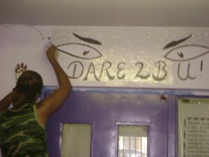 Person painting 'DARE 2 B' on a wall above a purple door.