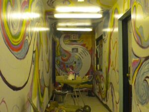 A narrow hallway with psychedelic murals and bright fluorescent lights.