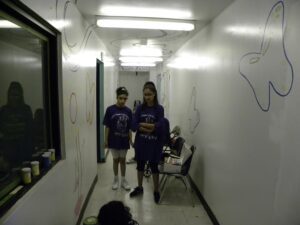 Two people in purple shirts standing in a white hallway with chairs.